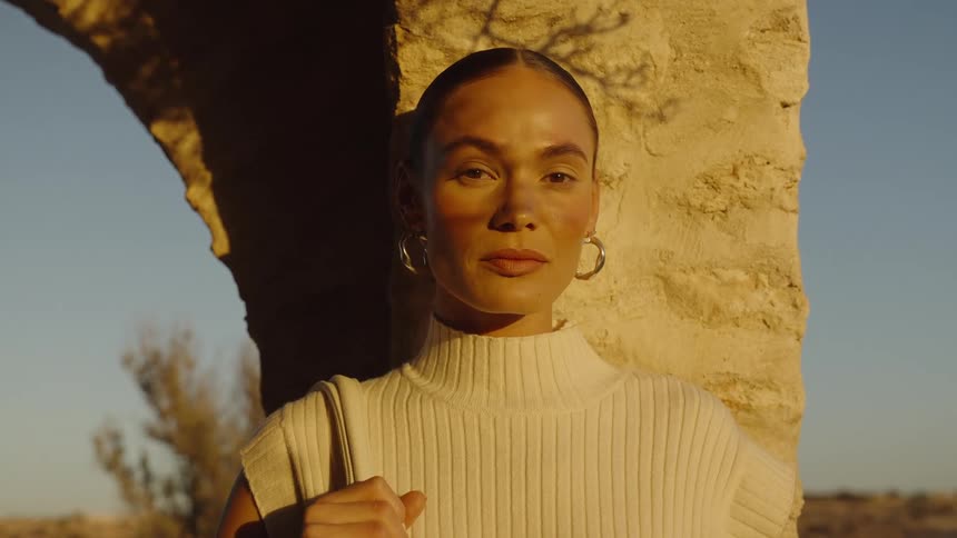 A close view shows a woman wearing a light colored knit top and hoop earrings standing framed by a sunlit rustic stone archway