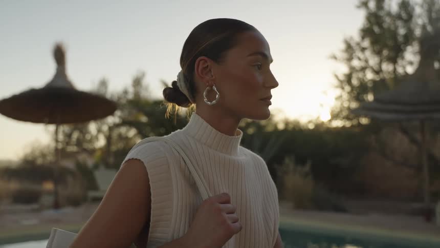 A close view shows a woman wearing a sleeveless knit top and hoop earrings turning from profile to face the camera by a pool at sunset