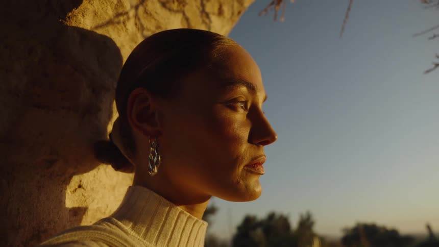 A close view of a woman with her hair pulled back and wearing hoop earrings and a white turtleneck standing near a stone archway in warm evening sunlight