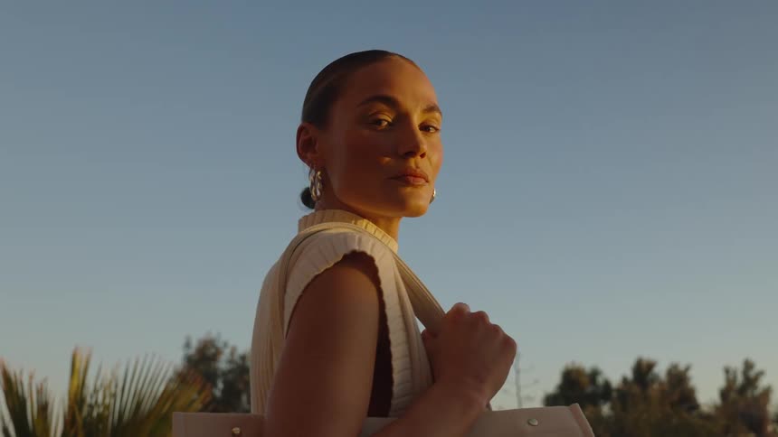 A close view shows a woman wearing a sleeveless knit top and hoop earrings standing outdoors with the golden light creating a strong outline on her profile
