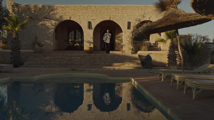 A woman carrying a large bag walks out of a house down the stairs toward a pool