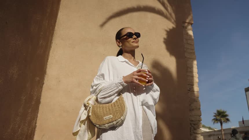 A woman in sunglasses and a loose white shirt stands outdoors in the sun possibly in a warm southern climate she is holding a glass of iced drink and carrying a small woven straw bag over her shoulder