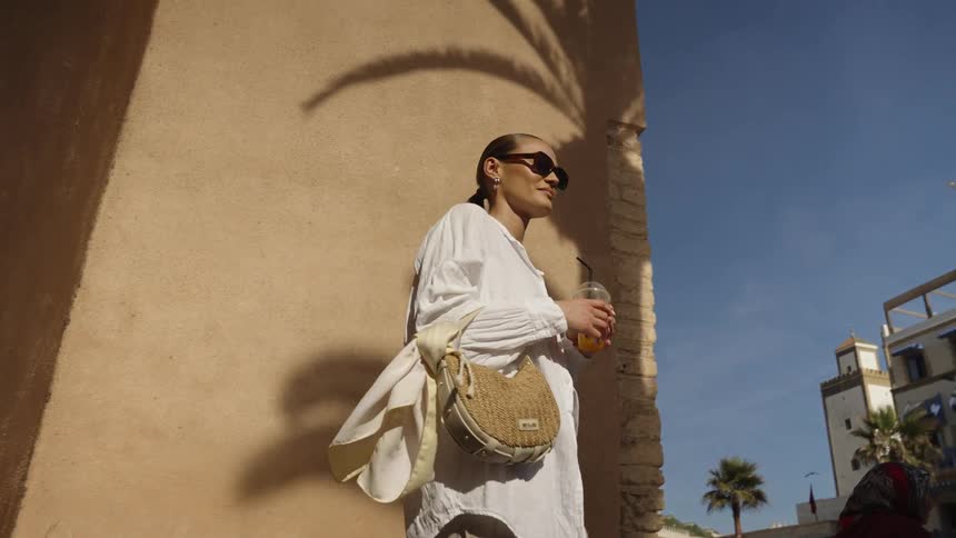 A fashionable woman in sunglasses and a white shirt walks outside along a sunlit tan wall holding a beverage and casting a palm tree shadow