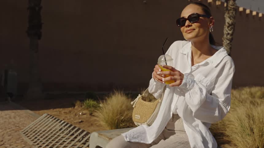 A woman walks to a spot holding a drink and sits down as she smiles