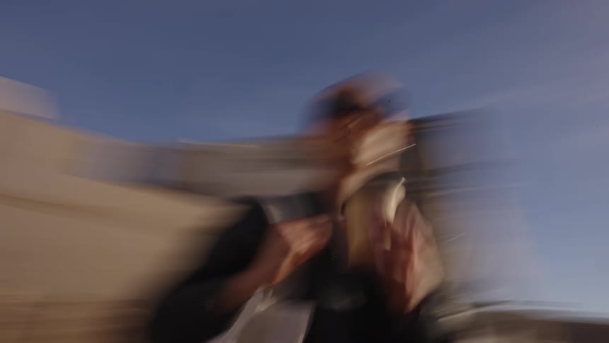 The video shows a quick shaky lowangle shot of a person with long dark hair wearing sunglasses and a lightcolored coat walking past a weathered whitewalled building they are carrying a drink in one hand and a large tote bag suggesting movement t