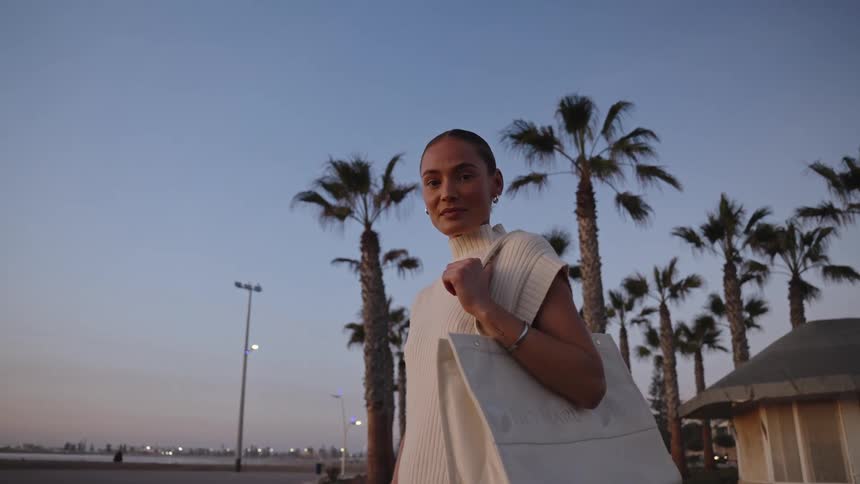 Woman at dusk holding a large bag on a palmlined promenade