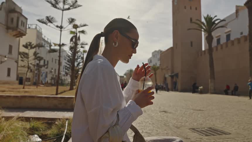 A woman wearing sunglasses and a white shirt is sitting outside in a sunny historic city square possibly in morocco or a similar mediterranean regionshe is taking a sip of an orange drink through a straw with a large ancient tower visible in the ba