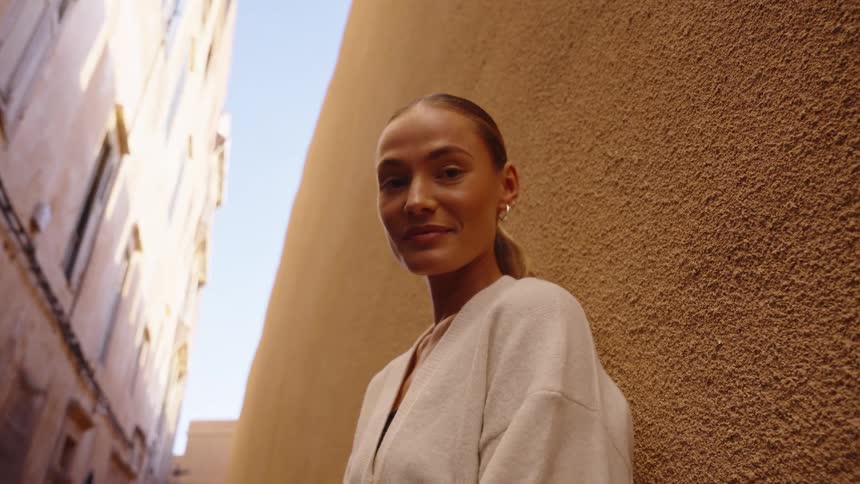 A close view shows a smiling woman wearing a light cardigan looking directly into the camera while leaning against a rough brown alley wall