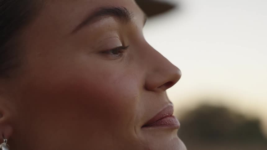 A young woman wearing makeup stands outdoors gazing ahead before briefly closing her eyes