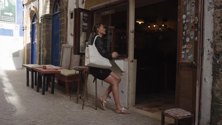 Woman sits on a stool at the entrance of a small caf in a narrow street