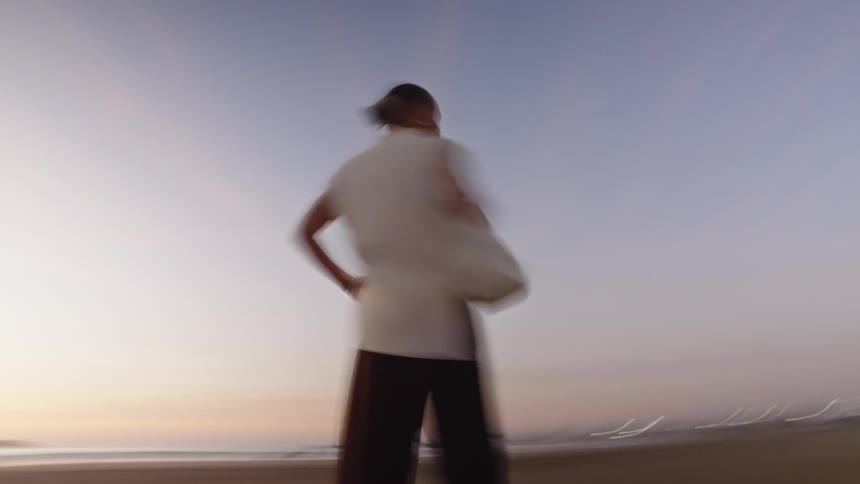 A blurry motionfilled shot captures a woman standing on the beach at twilight quickly spinning around to face the camera wearing wide dark trousers and a lightcolored top with the ocean horizon visible in the background