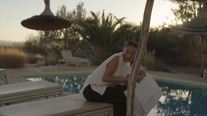 The video opens with a woman dressed in a white sleeveless top and dark trousers walking toward the camera alongside a sunny outdoor poolshe stops next to a lounge chair and sets down a large white tote bag then begins to look inside it while leaning
