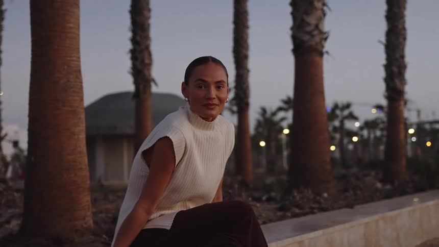 A woman relaxes on a low wall at dusk surrounded by palm trees and twinkling lights