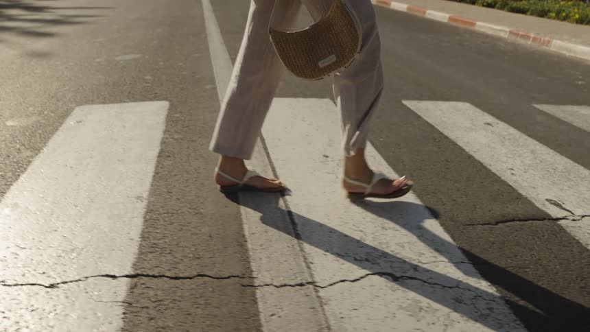 A woman wearing a white shirt and sunglasses walks confidently across a sunlit crosswalk carrying an orange drink and a woven bag on a warm city street