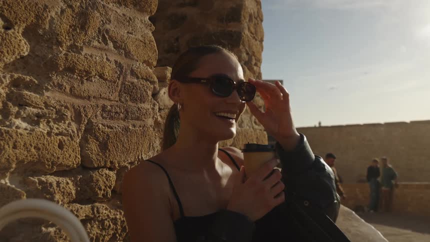 A young woman rests behind a brick wall on the street wearing a black jacket holding her sunglasses and a coffee cup smiling on a sunny day