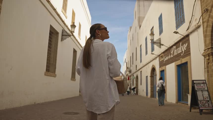 A woman wearing a white shirt and sunglasses walks down a narrow sunlit street between white and blue buildings carrying a woven bag