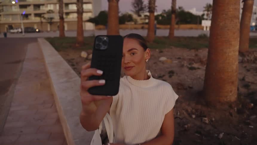 A close view shows a woman in a white top smiling while recording a selfie video with her phone on a promenade lined with palm trees at sunset