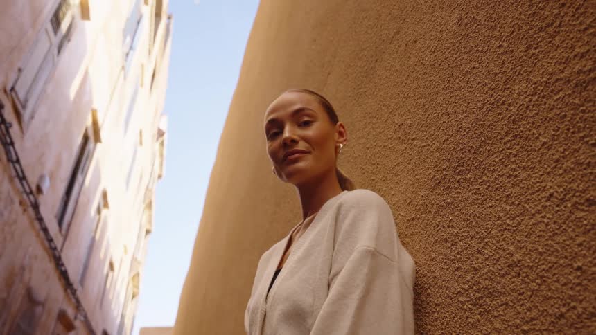 A close view shows a woman wearing a light cardigan standing against a textured brown wall in a narrow sunlit alleyway looking forward