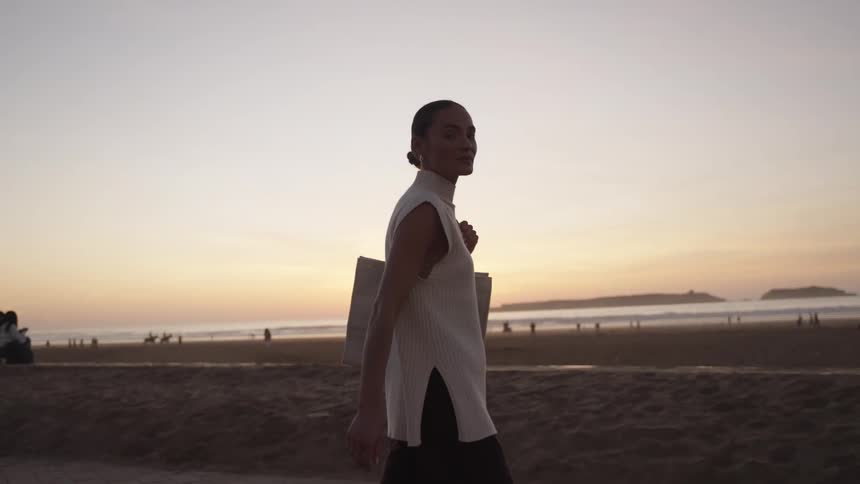 A close view shows a woman walking on a sandy beach at sunset highly silhouetted against the bright golden orange sky