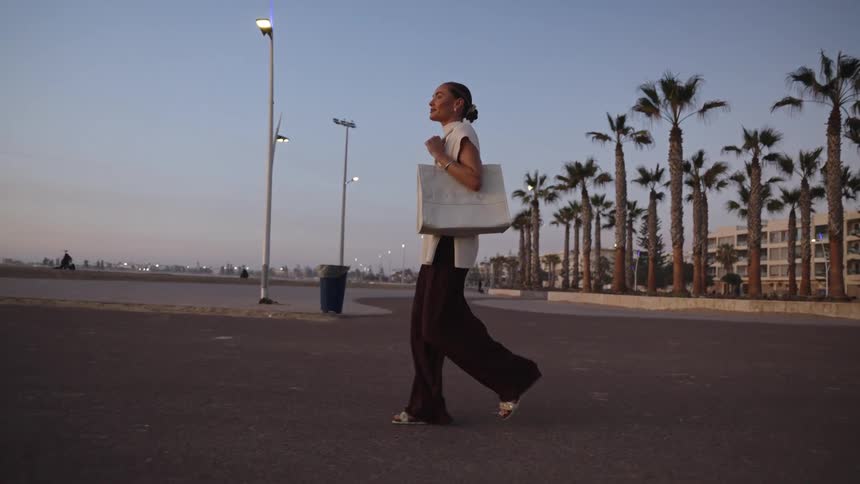 A stylish woman carrying a large white tote bag crosses a street at dusk near a beach promenade lined with palm trees
