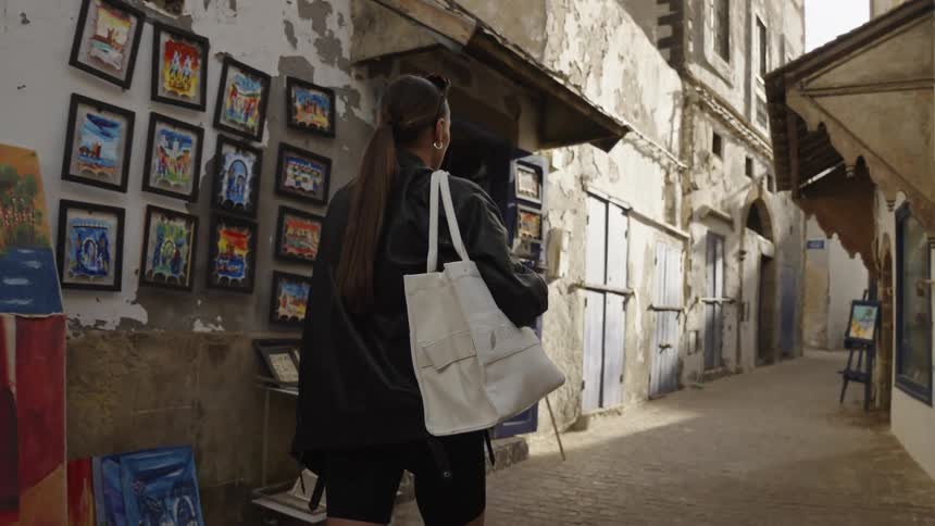 Woman walks along a narrow street observing paintings displayed on an old wall