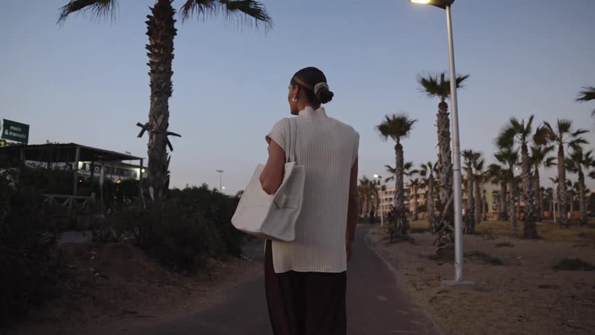Woman walking at dusk along palmlined path with white tote bag and restaurant in the background