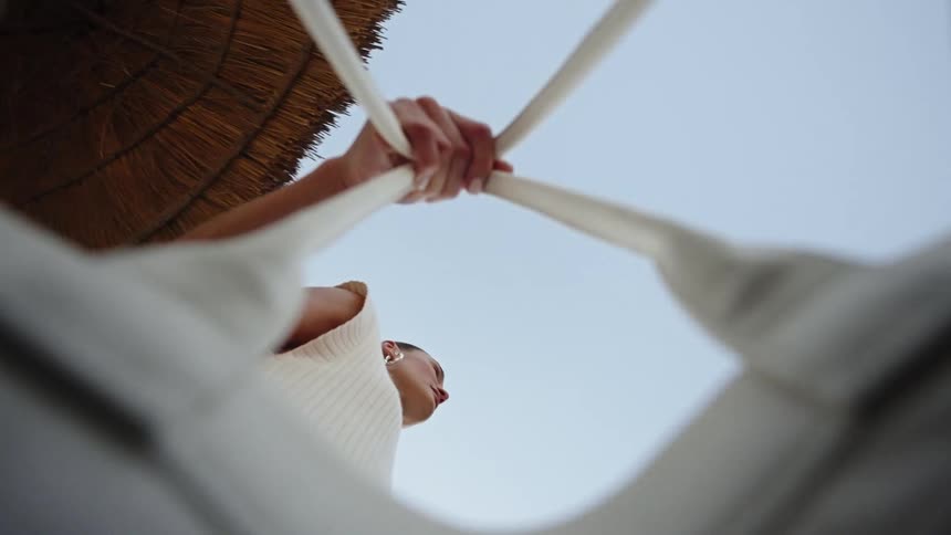 This video features an extreme lowangle shot looking up at a person lying down possibly in a hammock or hanging chair with a clear blue sky overhead the persons hands are gripping four converging white ropes or fabric straps above them