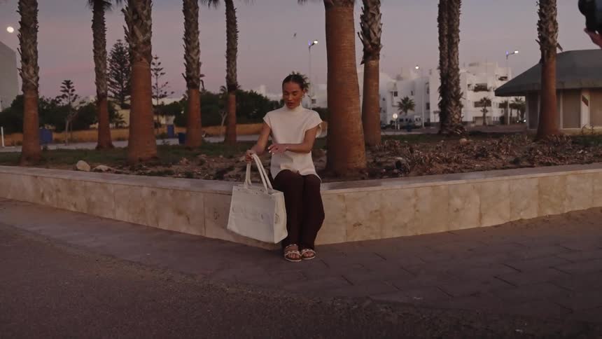 Woman holds white bag beside palm trees on urban walkway during twilight