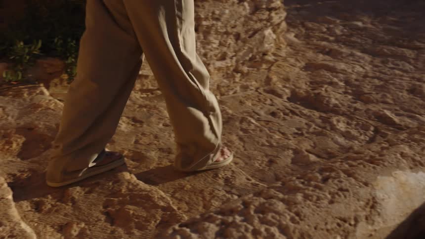 A lowangle shot focuses on the lower body of a woman wearing lightcolored widelegged trousers and sandals as shecarefully walk down a set of rough weathered and uneven stone steps bathed in warm sunlight