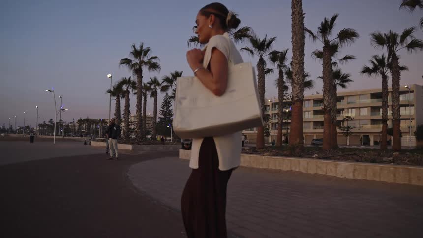 The video shows a woman with her hair pulled back in a bun wearing a white sleeveless top over dark pants and carrying a large white tote bag on her shoulder she is walking on a paved promenade lined with palm trees and modern residential buildings in