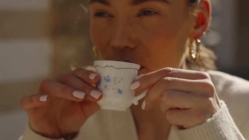 Closeup of a woman sipping coffee from a small patterned cup outdoors in bright sunlight