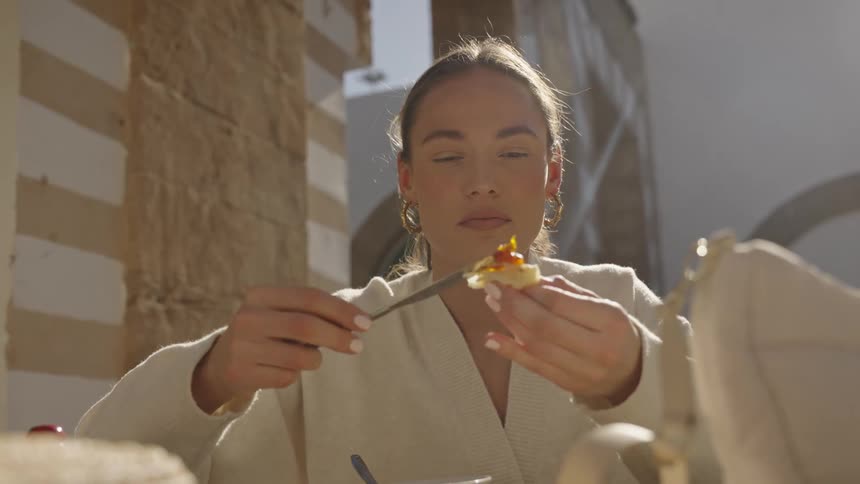 A close view shows a woman wearing a light robe spreading jam on a small piece of bread while eating breakfast outdoors in a sunny courtyard