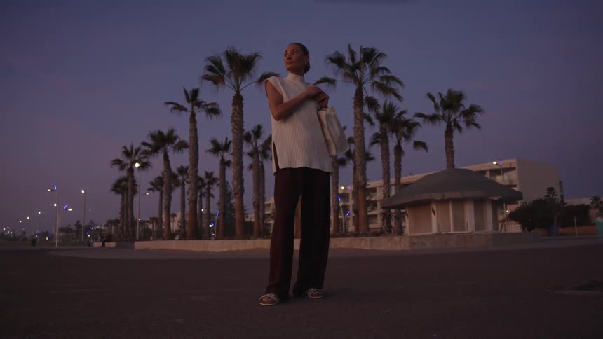 A woman with her hair pulled back stands in the center of a paved area holding a tote bag and gazing toward the left while wearing a white sleeveless top and dark wideleg pants set against a dark blue twilight sky and with tall palm trees
