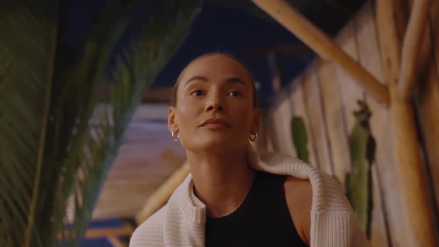A woman with her hair tied back is standing in a dimly lit tropicalstyle setting with wooden beams and foliage she is wearing a dark top with a white knitted cardigan draped over her shoulders and she looks up and smiles