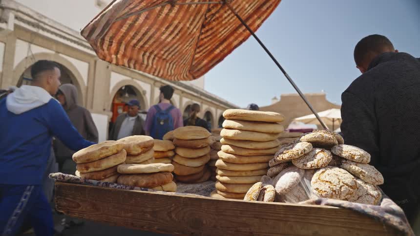 A vendor on a street in a market is selling bread as people walk past
