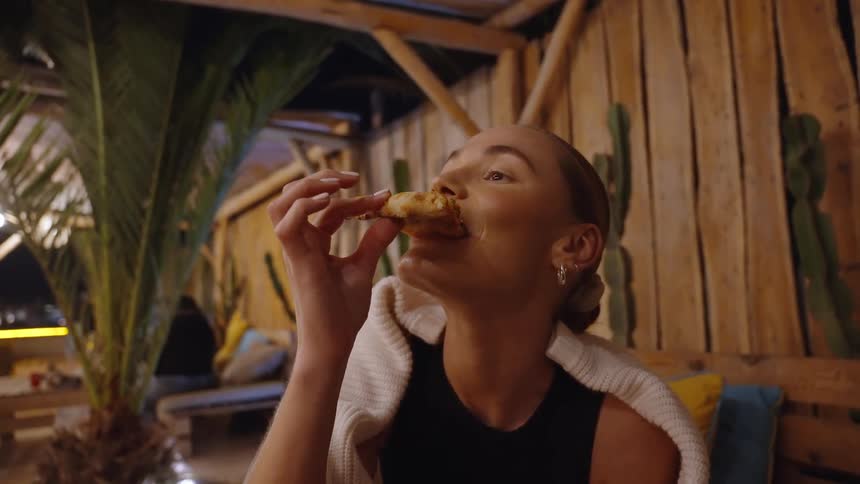 A woman eats a slice of pizza while sitting outdoors at a rustic restaurant