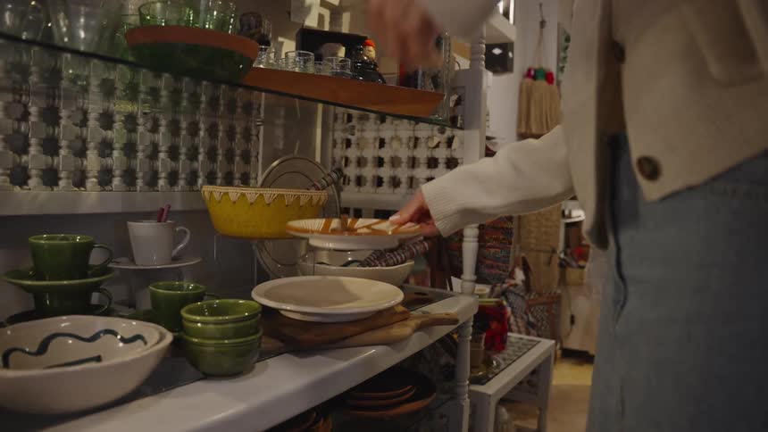 A woman picks up a single plate with a wooden spoon from a stack on a shelf filled with ceramic dishware