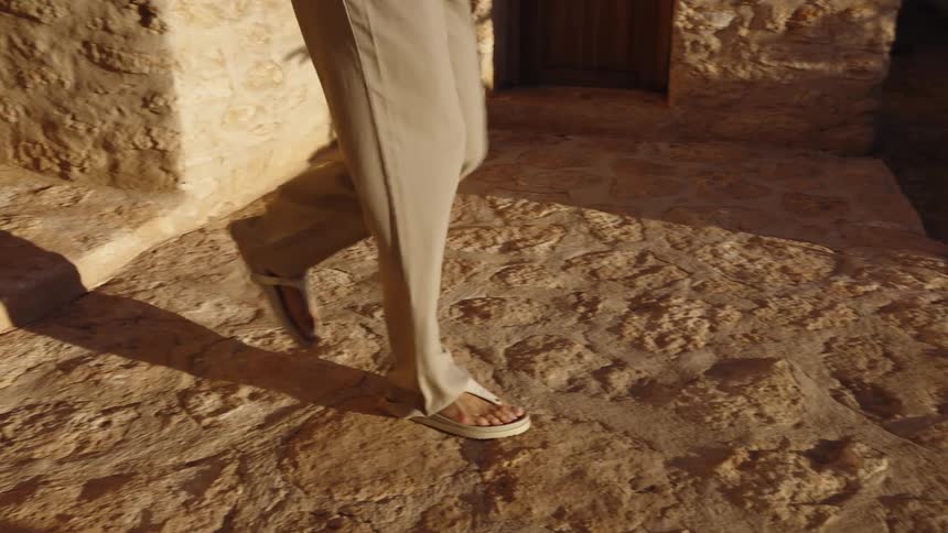 A person wearing lightcolored trousers with a slit at the ankle and white thong sandals walks down rough stone steps and a path the setting appears to be an old sundrenched location with stone walls and wooden doors