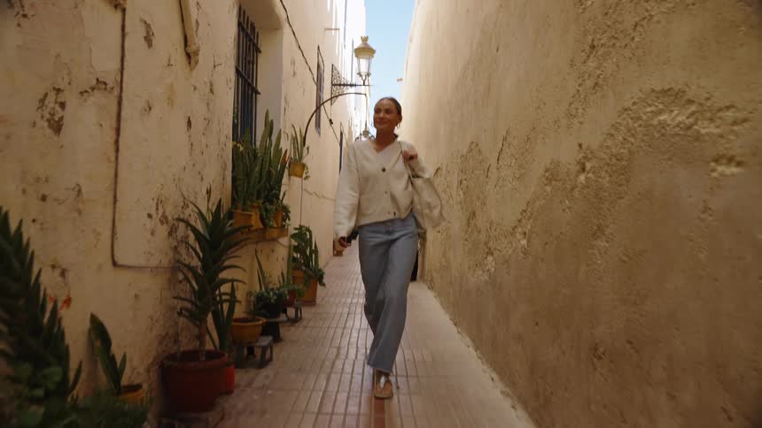 The video features a smiling young woman walking confidently down a very narrow highwalled alleyway possibly in a mediterranean or north african townthe walls are old and textured with a row of potted succulents and cacti lining the left side add