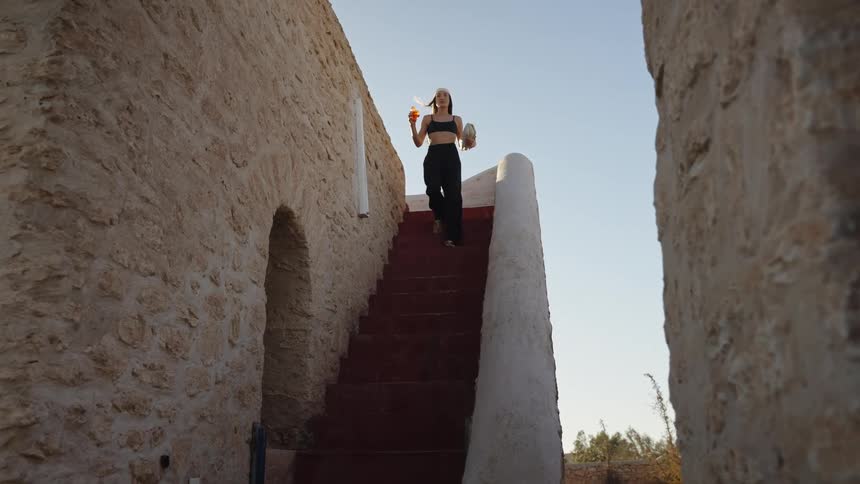 The wind blows a womans hair around as she walks down a staircase with a drink and books