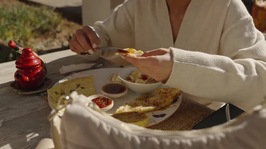 A woman wearing a white cardigan sits at an outdoor table and spreads jam onto a piece of bread eating a meal in bright sunlight