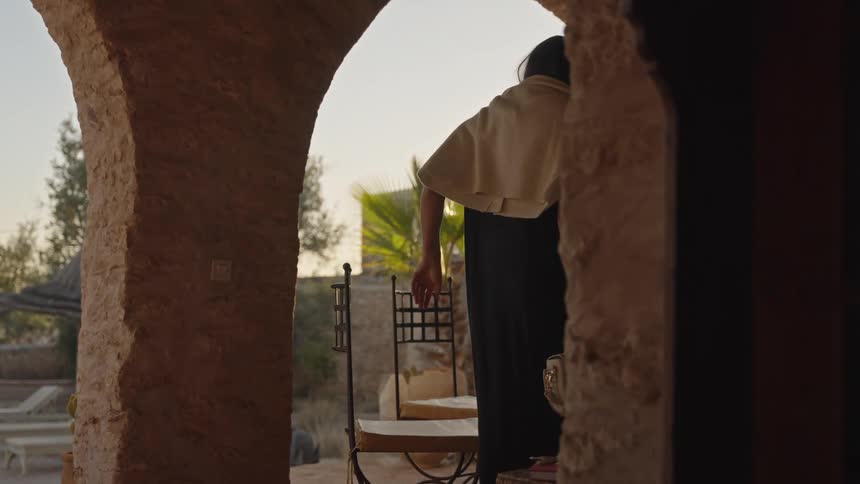 A close view shows a woman in a dark dress and shawl walking through a stone arch and sitting down at a patio table near a sunlit pool