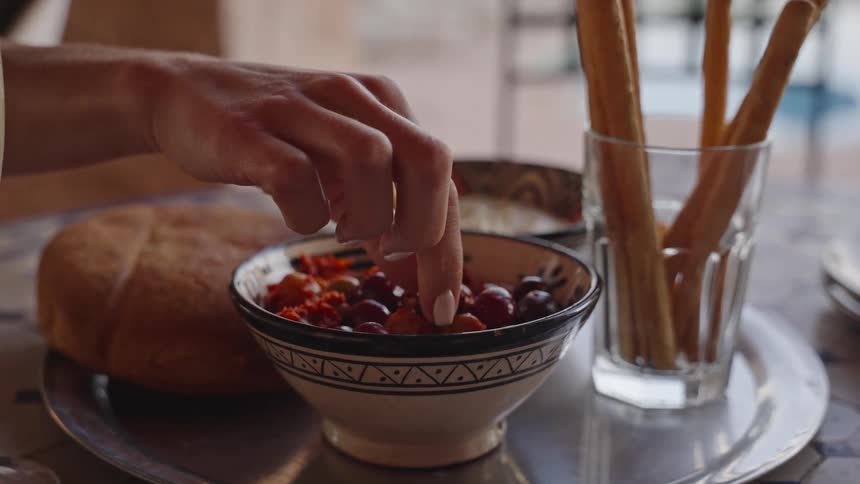 A persons hand reaching into a small ceramic bowl filled with olives and other appetizers picking one out as a loaf of bread and a glass of breadsticks sit on a tray nearby