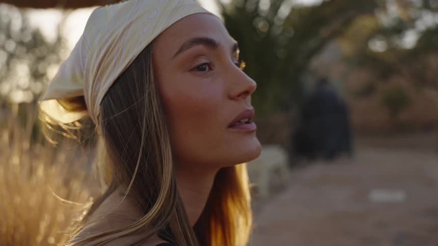 A young woman wearing a lightcolored bandana is shown in profile looking offcamera she is outdoors possibly near a pool with warm golden light illuminating her hair from behind