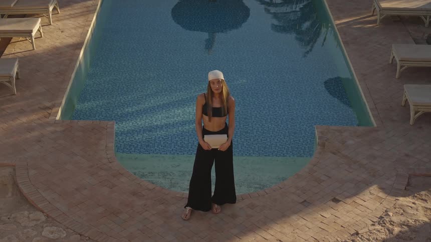 This video features a highangle fullbody shot of a young woman standing at the edge of a curved tiled swimming pool she is wearing dark wideleg pants a dark top and a lightcolored beanie holding a small clutch or book in front of her