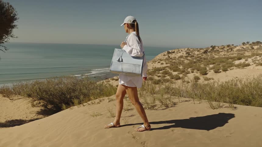 A close view shows a woman wearing all white and a cap walking across a bright sand dune ridge above the sparkling blue ocean