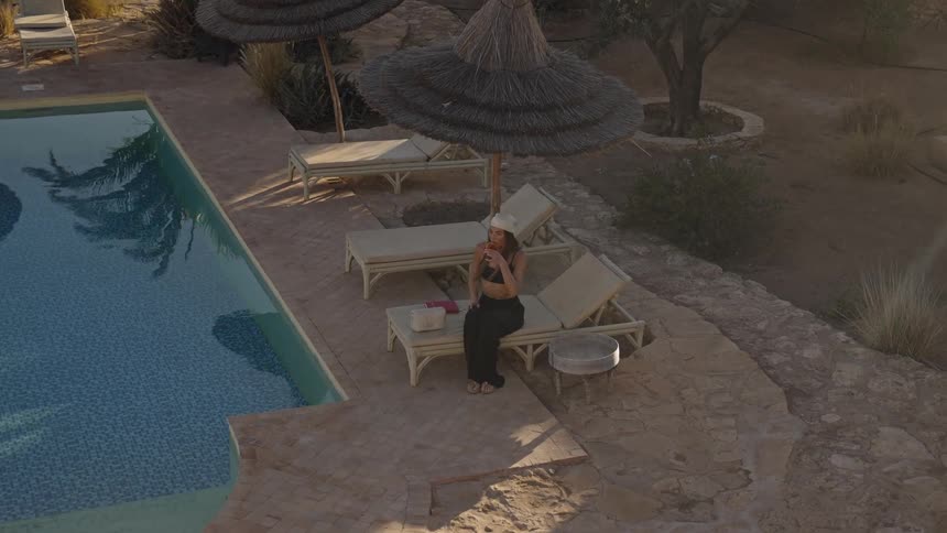 A woman relaxes on a lounge chair next to a pool enjoying a drink in a resort