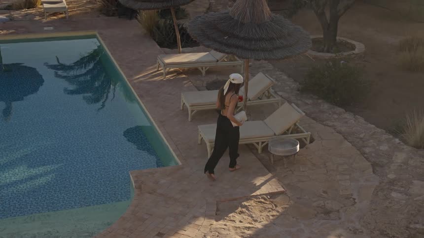 An overhead view shows a woman in dark trousers and a white cap walking beside a tiled swimming pool and setting down a drink on a lounge chair table
