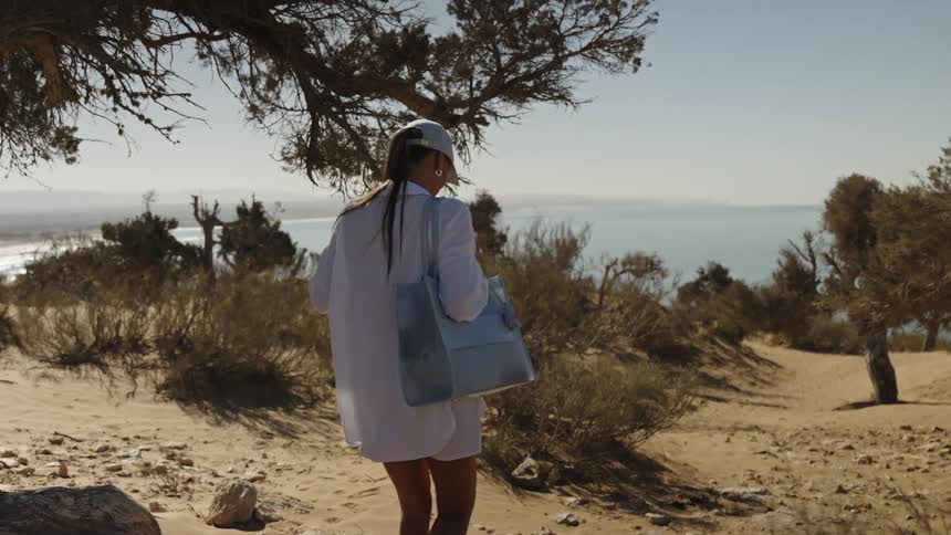 A woman wearing white clothes and a cap as she walks down a sandy path on a small hill moving toward a beautiful view of the bright blue ocean visible in the distance