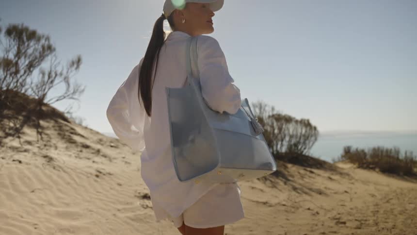 A woman wearing all white including a longsleeve shirt shorts and a baseball cap walks across a sandy dune under bright sunlight carrying a large lightblue tote bag over her shoulder the background shows scrub brush a gnarled tree and a distant